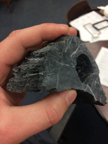 A chunk of dark rock. There is a hole visible on one side where a core was drilled from the rock. The sample is fist-sized.