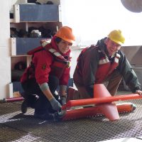 Students wearing hardhats prepare a bright colored instrument on the ship's deck.