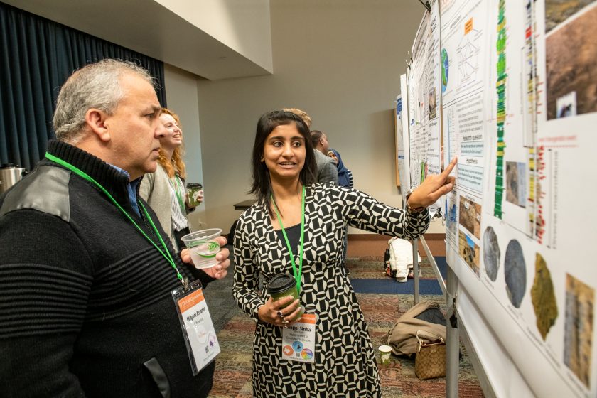 Sinjini points at her poster as Miguel intentlyl istens.