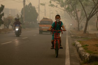 Child wearing a mask on a red bicycle. Cars nearby are dimly visible through thick haze