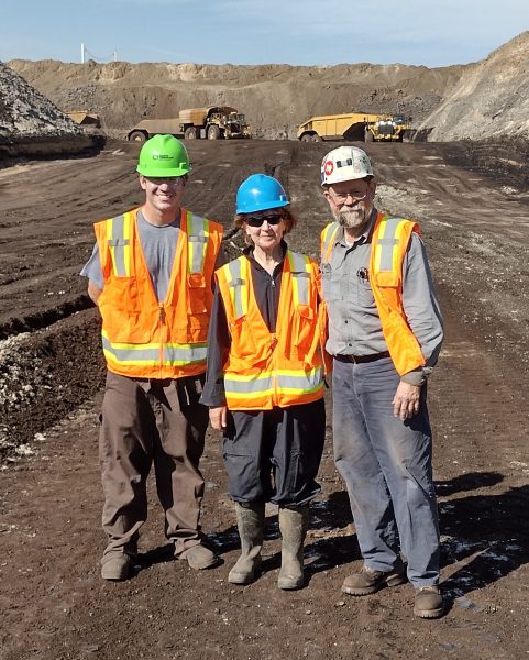 Three people in safety vests and hard hats stand in a coal mine with heavy machinery in the background.