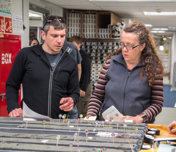 Photo of two researchers standing behind a table on which split cores are laid out. Parts of the core are labelled. From their surroundings it is clear they are on a science vessel.