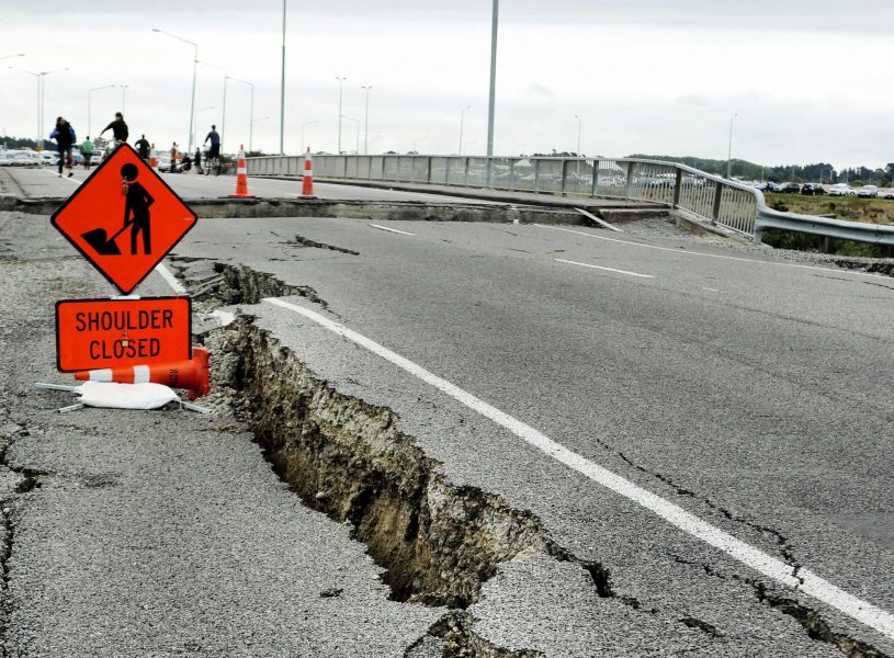 Photo of a large break in a road, the right side is about a foot higher than the left. A road sign reads 'Shoulder Closed' and there are road workers some distance away where another break is visible crossing the road width.