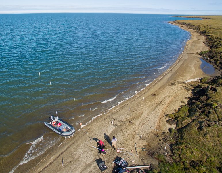 A large swath of blue water meets the sandy shore of northern Alaska in an aerial photograph. A boat and researchers with their instruments are in the foreground.