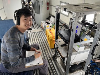 Picture of Kristian wearing headphones sitting at a control panel holding a clipboard. He is on an aircraft.