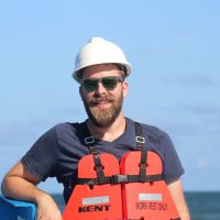 Portrait photo of Chris wearing a hard hat and life vest with the ocean behind him.