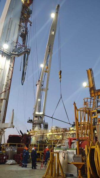 A crane towers over a rig deck. Blue clad rig workers are below. The crane is carrying a long core tube horizontally.