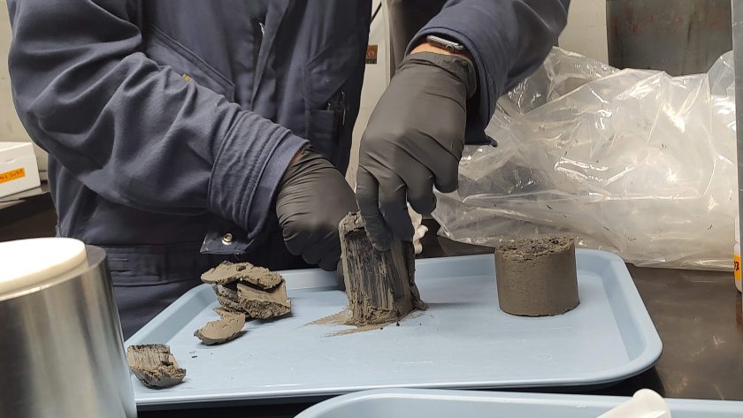 Close up of a scientists slicing into segments of core like a cake. The core is in cylindrical chunks a few inches tall and is dark grey.