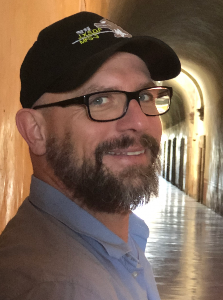 Portrait photo of Thorsten Becker in an arched corridor, wearing a cap.