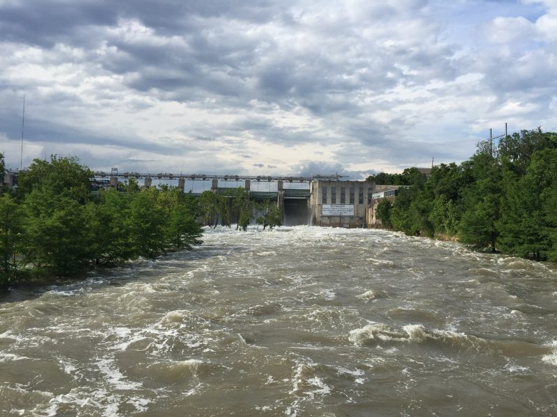 Picture of the Colorado River facing the dam. The waters are brown and choppy and as high as the treeline on either bank.