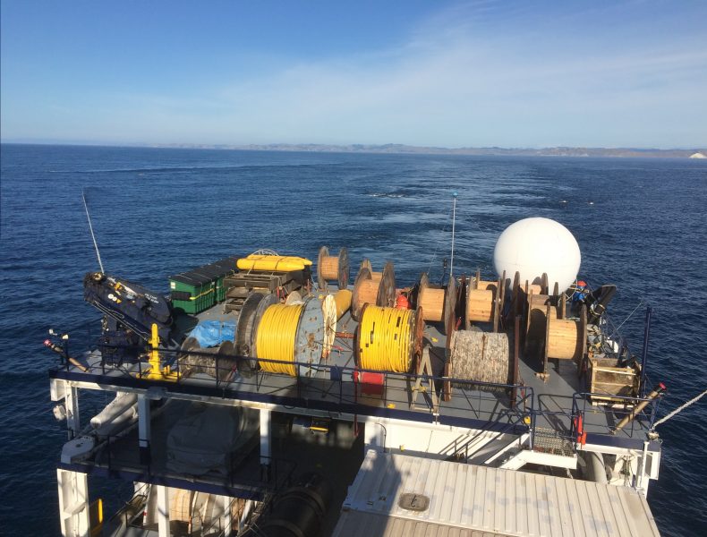 Photo from a high vantage of instruments on the aft of a ship. Just visible in the water in the ship's wake are wave crests from the seismic streamer towed by the ship, visible just under the sea surface.