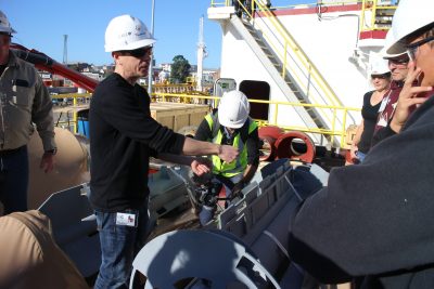 Photo of Demian wearing a hard hat and standing over an instrument on the deck of a ship and talking to other scientists, also in hard hats.