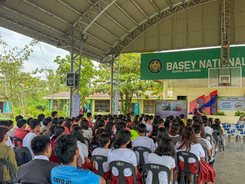 A large group of students sit under a large, rounded metal roof while Cardenas lectures.