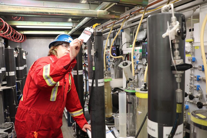 Phillips is wearing red protective gear and hard hat and is examining one of the upright pressure cores in a lab.