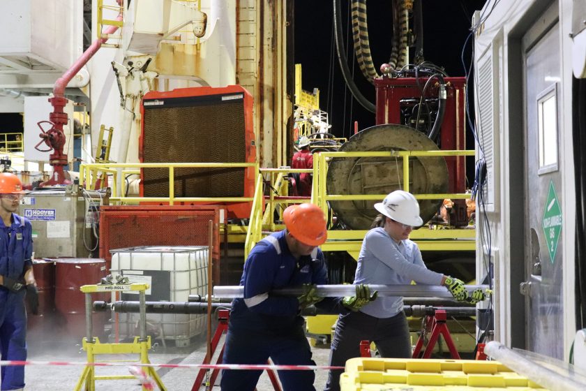 Two people pull on a long clear plastic pipe (the lining) that runs into a nearby building. It is night time and they are surrounded by heavy machinery.