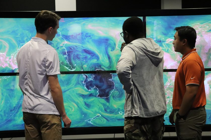 Three students, with their backs to the camera, look at a large screen that shows swirls of blue and green colors over a map of the world.