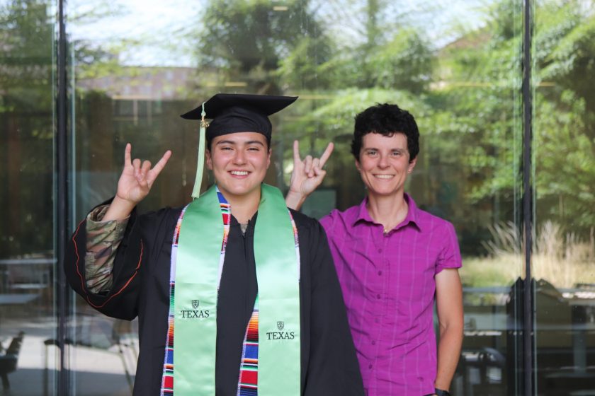 A female student in a cap, gown and graduation stole stands with her female professor in a fuchsia collared shirt giving the UT hook em sign with their hands. 