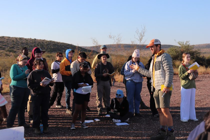 A professor in the foreground gestures while students behind him write notes in their notebooks. They're outdoors in a desert landscape.