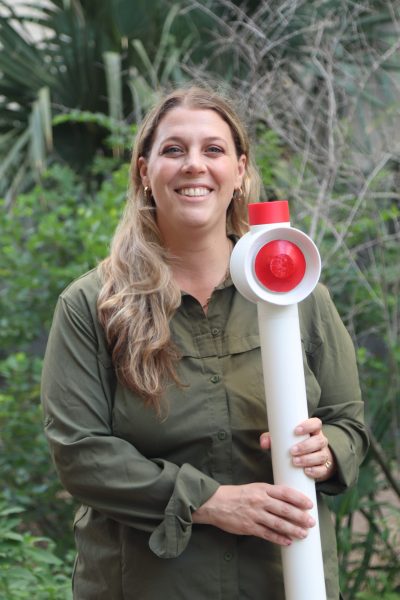 A woman smiles and holds up a 3D-printed model of a flood alarm--a white tube with a circular red alarm at the top.