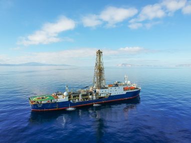 The ship at sea as if looked by a passing bird. The ship’s drilling riser is above the horizon, where islands are visible in the distance.