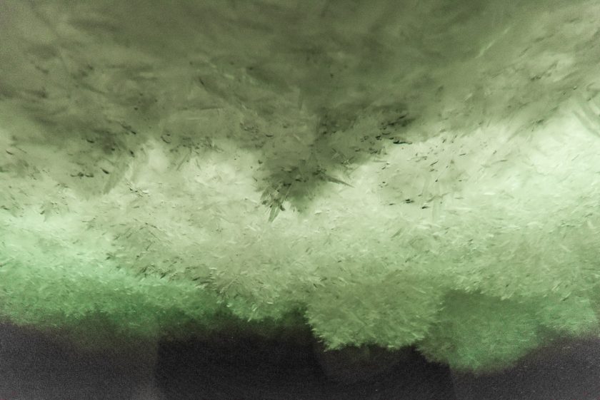 A view under an ice shelf with mounds of pale green snow-like ice suspended over dark depths.