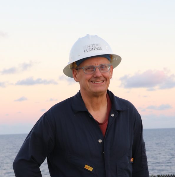 Peter Flemings smiles against the backdrop of the Gulf of Mexico at sunrise. He wears a white hard hat and dark blue coveralls.