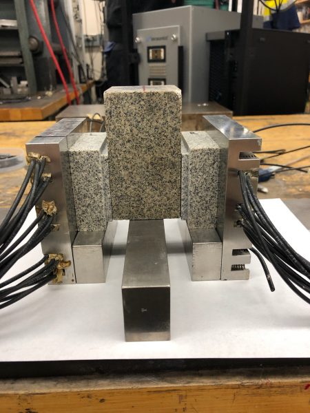 Photo of three granite-like blocks in an instrument that looks like a metal vice, connected to thick heavy cables. 