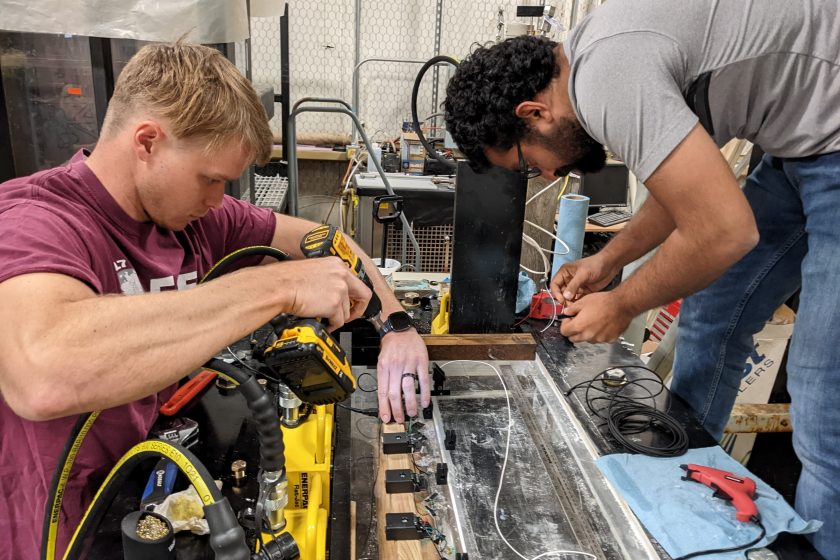 Two people working with a drill and glue gun over a wire-covered table-like device in a workshop-like environment.