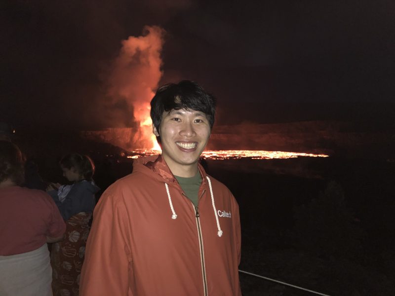 A man smiles in front of an erupting volcano in the distance, at night
