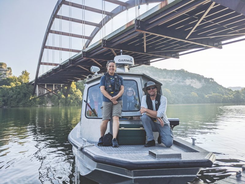 The crew pose on the deck and inside the boat. The bridge is visible behind them.