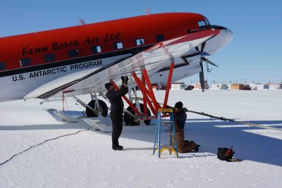 Picture showing a propeller plane parked on ice, with engineers working under the wing