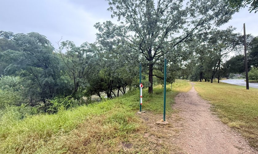 The flood monitor device is shown mounted against a pole along a walking path, with a river in the background.