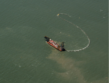 An aerial view of a dredging vessel at sea. Trails of sand are visible in vessel's wake.