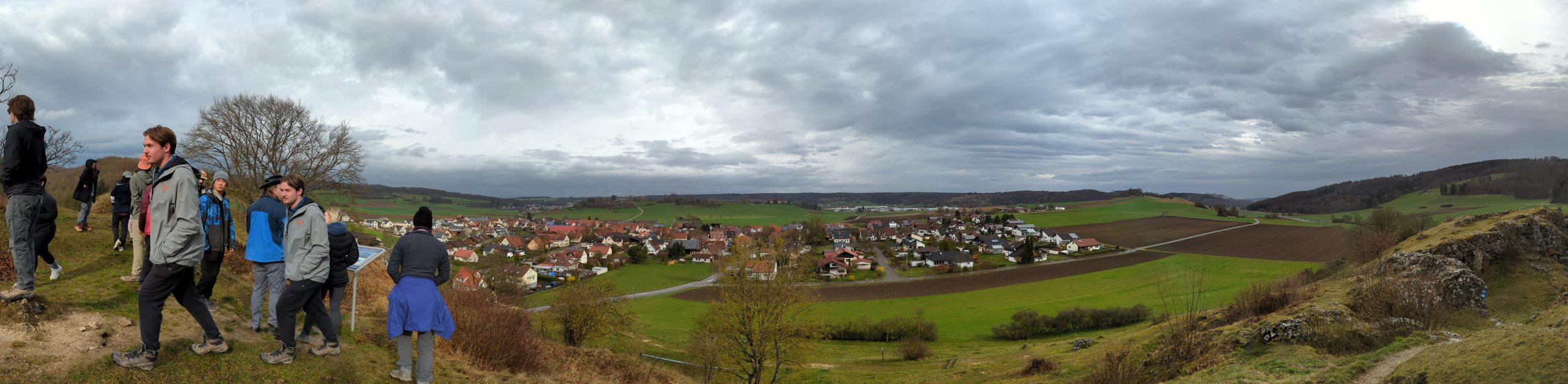 A view from a high point across a small German village and surrounding green farmer's fields. The sky is overcast (grey and cloudy).