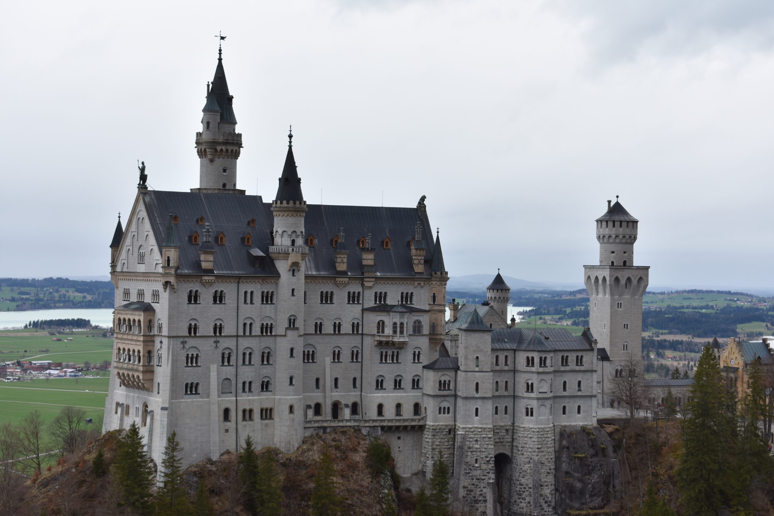 A view of Neuschwanstein castle from afar. It's a white, German-style castle that's approximately 7 stories high, with grey roof tiles and tall, thin spires. The castle is perched upon a hill.