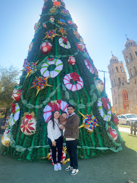 The author, Katherine Garcia, with her fiance and baby son, outdoors in front of a giant decorative christmas tree.