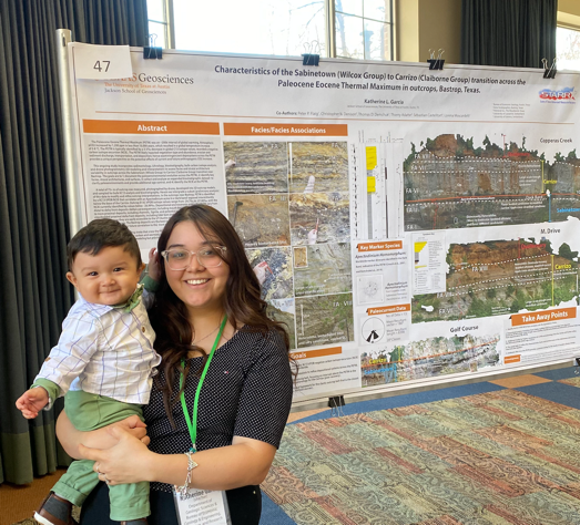 Katherine holding her baby son at the 2023 Poster Symposium, standing in front of a poster about her research.